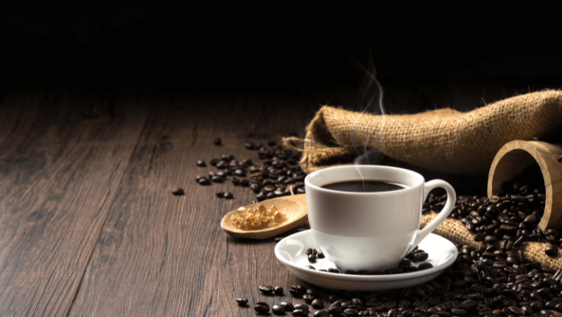 Hot coffee in a white coffee cup and many coffee beans placed around and sugar on a wooden table in a warm light atmosphere, on dark background, with copy space